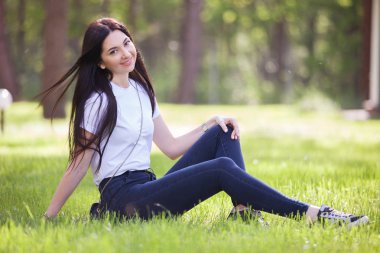 Genç kadın yeşil çimlerin üzerinde parkta dinleniyor. Renkli arka planlı güzel doğa sahnesi, yaz mevsiminde ağaçlar. Açık hava yaşam tarzı. Mutlu gülümseyen kadın yeşil çimlerde oturuyor.