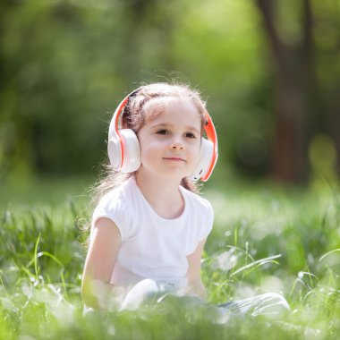Şirin küçük kız parkta müzik dinliyor. Aile açık hava yaşam tarzı. Yeşil çimlerin üzerinde oturan kulaklıklı mutlu küçük çocuk. Yazın güzellik doğası. Çocukluk mutluluğu. Çocuk günü.