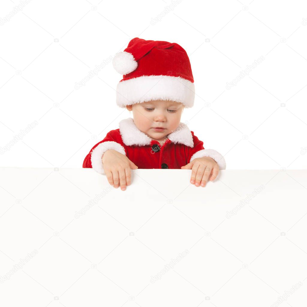 Happy santa baby with white blank board isolated on white. Small model in santa hat at studio holding paper poster. Christmas, xmas, winter concept. Santa girl looking at white blank board