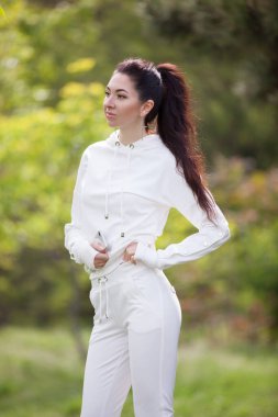 Beyaz kostümlü, parkta yürüyen hoş bir kadın. Renkli arka planlı güzel doğa sahnesi, yaz mevsiminde ağaçlar. Açık hava yaşam tarzı. Moda biçimi