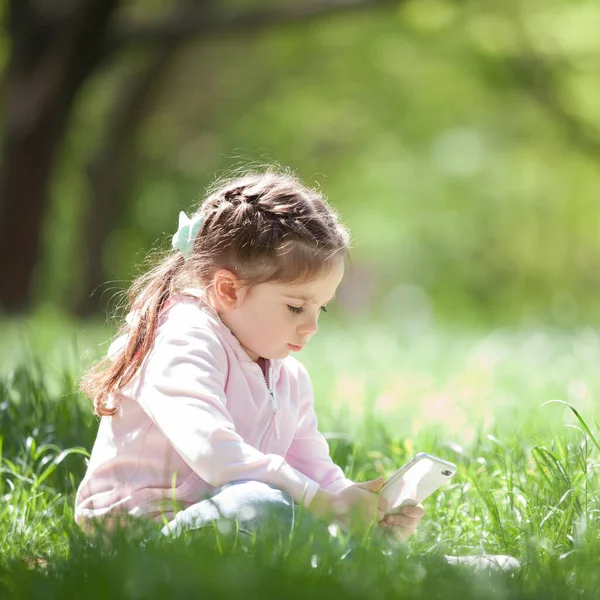 公園で携帯電話を持つかわいい女の子 家族のアウトドアライフスタイル 幸せな小さな緑の草の上に座っている 夏や春の美しさ 子供の頃の幸せ モバイル チルドレン — ストック写真