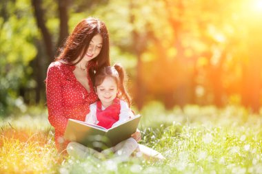 Mutlu anne ve kızı yaz parkında bir kitap okudu. Aile hayatı olan güzel doğa manzarası. Mutlu aile yeşil çimenlerde dinleniyor, sarılıyor ve dışarıda eğleniyor.