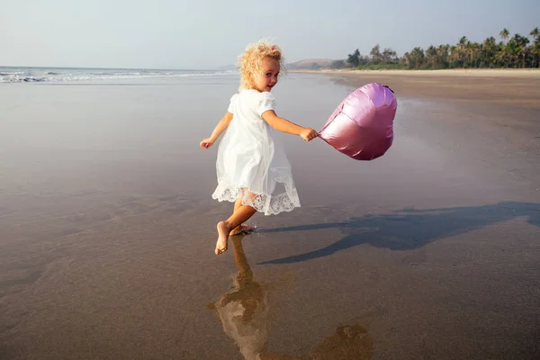 Маленькая кудрявая девочка с розовыми шариками на пляже на закате — стоковое фото
