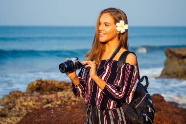 tropikal zevk yaz turizmi ve fotoğrafçılık. Deniz plajı hint okyanusu goa turkuaz su, gökyüzü ve kayalar yakınında kamera ve sırt çantası ile şık bir çizgili tulum genç seyahat kadın