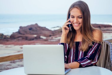 Tropik cennet plajda dizüstü bilgisayar ve telefon freelancing kullanarak şık çizgili tulum genç seksi kadın. Kız serbest çalışma bir yaz kafe de oturan uzaktan iş hint okyanusu arka plan