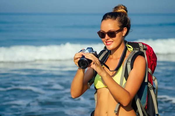 Freiheit junge Backpacker in einem sexy gelben Bikini-Badeanzug hält eine Kamera in der Hand an einem Strand. Blonde Fotografin Frau steht in der Nähe des Meeres mit großen Touristentasche auf den Schultern, Tourismus Meer — Stockfoto