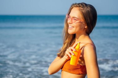 Hindistan Okyanusu kıyılarında mavi denize karşı sprey spf güneş bloğu bir şişe tutarak tabaklanmış kol ve omuzlar üzerinde güneş koruması uygulayan şık bir mercan turuncu bikini mayo kadın