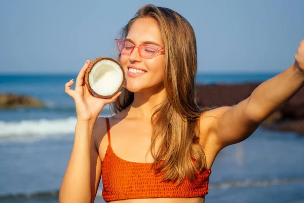 happy smiling girl in stylish orange coral swimsuit taking selfie on phone with coconut in vacation.bikini woman holding cellphone photo camera smartphone pictures on the ocean india.spa coco nut oil