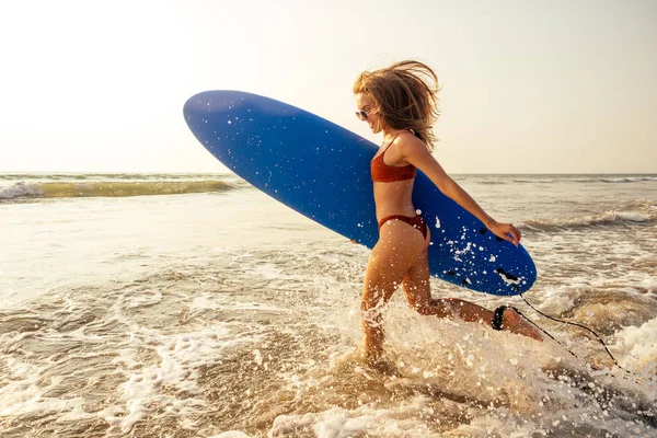 Surfista ragazza con tavola da surf blu sulla spiaggia di sabbia. sexy modello femminile bodybuilding e fitness in un bikini arancione costume da bagno giovane donna bordo al paradiso tropicale . — Foto Stock