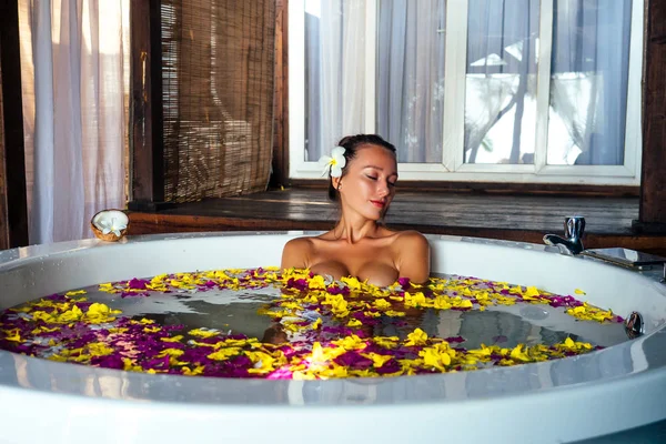 Hermosa chica morena con plomería en su pelo en jacuzzi con pétalos de rosa y flores tropicales. Cuidado del cuerpo, luz natural maquillaje peinado y relaxation.sexy hermosa mujer en un día de baño para mujer — Foto de Stock