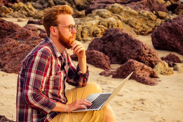 Hombre freelancer barbudo gafas hipster con un ordenador portátil de trabajo remoto en la playa en la puesta de sol.freelancing chico que trabaja cerca de las rocas y el hombre sea.business de vacaciones en un paraíso tropical del desierto — Foto de Stock