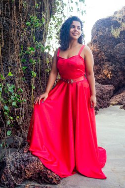 Curly-haired young afro american girl in a red long silk dress on a corset on the background of tropical rocks and vines.Female dark skin model indian woman enjoing at the sea coast paradise beach