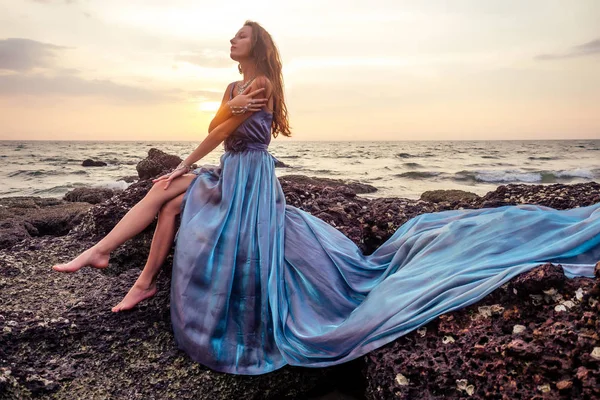 gorgeous style brunette young woman in blue chameleon dress long train standing on a rock near the sea, sand tropical on a rock.beautiful mermaid lonely melancholy waiting freedom and freelancing