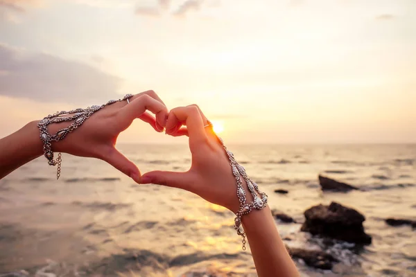 Frauenhände in Herzform gegen den Himmel ziehen an Sonnenstrahlen vorbei. Zeiger in einem luxuriösen Doppel-Silberschmuck Armband und Ring East-Stil in Form von Liebe Herz indischen Ozean Sonnenuntergang Valentinstag — Stockfoto