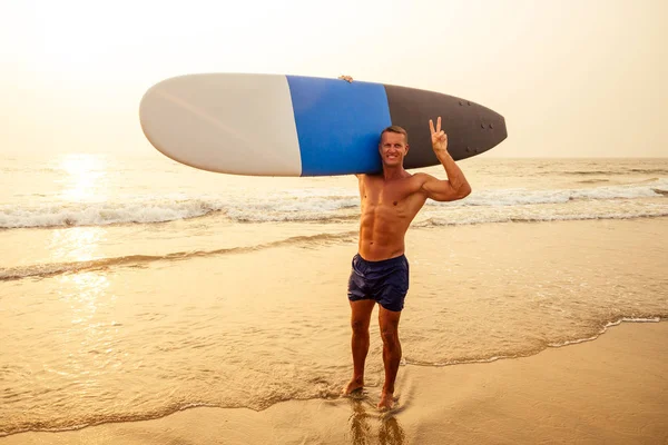 Knappe sportieve man die zijn surfplank op het hoofd draagt. Surfer tijdens de zomer reizen strand vakantie. Kaukasische sexy atleet fitness model dragen surfen surfplank. — Stockfoto
