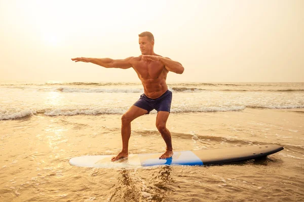 Surfer fitness man met een gespierd lichaam met zijn surfplank op het strand opwarmen. — Stockfoto