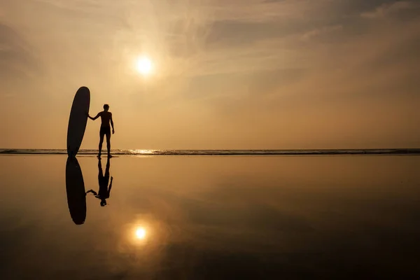 Een surfer man met zijn surfplank gaan om te voldoen aan de zee golven avond zonsondergang op het strand. mannelijke freelancer surfen op vakantie in een tropisch land Paradise Island — Stockfoto