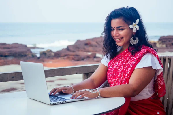 Remote working concept dream job.beautiful and young indian woman in red stylish sari, curly hairstyle, toothy white smile and flower in hair working with a laptop while sitting business woman — стоковое фото