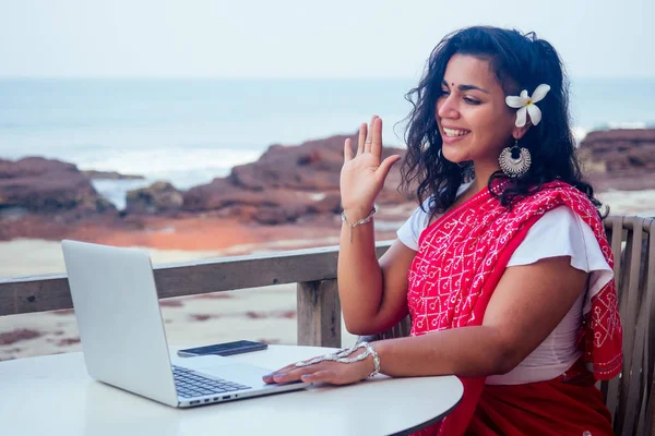 Concepto de trabajo remoto trabajo de ensueño hermosa y joven mujer india en rojo elegante sari, peinado rizado, sonrisa blanca dentada y flor en el pelo que trabaja con un ordenador portátil mientras está sentado en un café de verano por mar —  Fotos de Stock