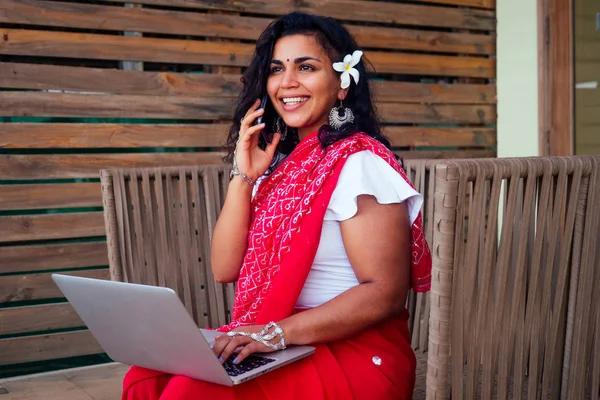 Atractivo india asiático mujer freelancer sentado en café. mujer en rojo indio elegante saree sari trabajando en el ordenador portátil y sonriendo hablando en el trabajo smartphone.remote sueño trabajo en la costa del paraíso —  Fotos de Stock