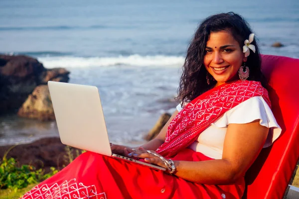 beautiful and young indian business woman in traditional india sari working with laptop by the sea.asian student girl remote work freelancer freelancing sitting on the beach using a smartphone phone