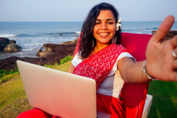 Remote working concept dream job.beautiful and young indian woman in red stylish sari, curly hairstyle, toothy white smile and flower in hair working with laptop photographing portrait selfie on phone — стоковое фото