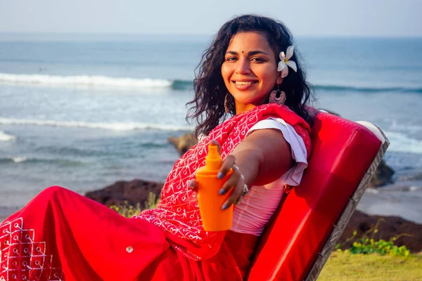 Retrato de um belo e sorridente sorriso branco-neve mulher indiana cabelo encaracolado preto e pele escura em um sari vermelho segurando garrafa de spray protetor solar em beach.girl desfrutando spf corpo paraíso férias — Fotografia de Stock