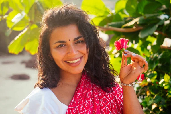 Indisches weibliches Modemodel mit weißem Zahnlächeln und Blume im lockigen Haar im traditionellen indischen Outfit roter Hochzeitssari, der in der Nähe tropischer Bäume an einem paradiesischen Strand am Meer posiert. — Stockfoto