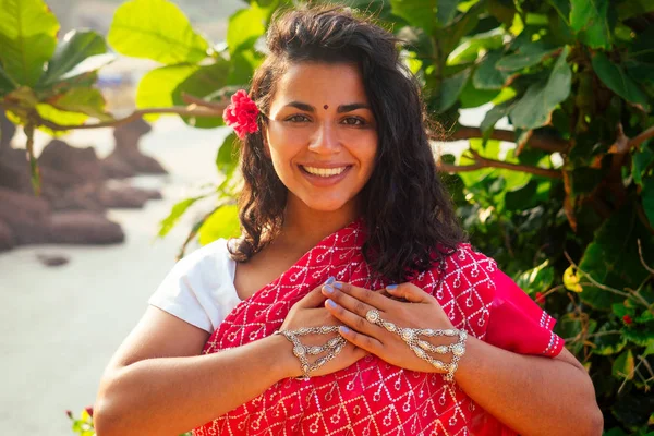 Indisches weibliches Modemodel mit weißem Zahnlächeln und Blume im lockigen Haar im traditionellen indischen Outfit roter Hochzeitssari, der in der Nähe tropischer Bäume an einem paradiesischen Strand am Meer posiert. — Stockfoto