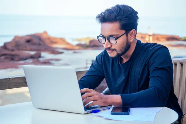 Schöner und erfolgreicher indischer Mann in einem stilvollen gut gekleideten Freiberufler, der mit einem Laptop am Strand arbeitet.Freiberufler und Fernarbeit.Geschäftsmann Student in einem Sommercafé am Ufer des indischen Ozeans — Stockfoto