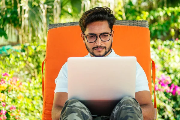 Remote work and freelancing.young indian man sitting orange lounger deck chair with laptop on beach.freelancer surfing the internet.freelance dark skinned stylish guy summer vacation tropical paradise — Stock Photo, Image