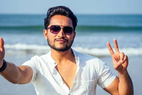 handsome and young indian male model beard and black hair haircut,fashion earring and glasses on a background of tropical ocean.selfie portrait man businessman student relaxing on the beach sea
