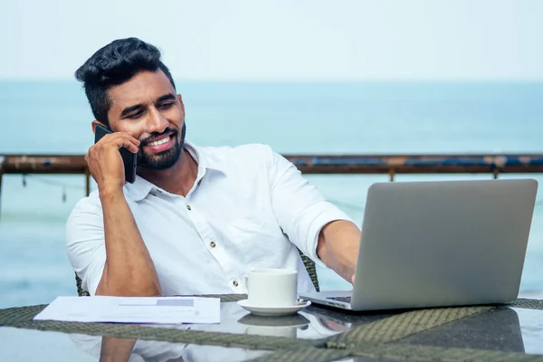 Красивий і успішний індійський чоловік у стильному добре одягненому фрілансеру працює ноутбук на пляжі.фрілансер і віддалена робота.бізнесмен студент в літній чашці чаю на березі Індії океану — стокове фото