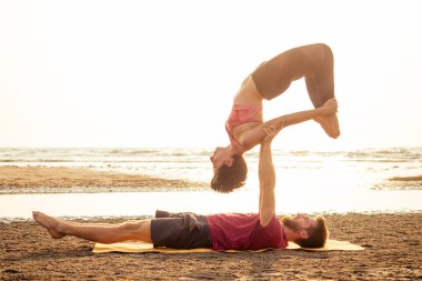 Young sportive couple girl and boy are practicing acroyoga exercises in the sunset on the beach clipart