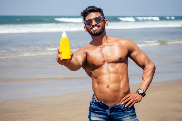 Happy young african man on the beach.Handsome and confident Masculine hard fitness bodybuilder with sixpack.India model male perfect abs, shoulders, biceps, triceps, chest holding a bottle of sunscreen — стоковое фото