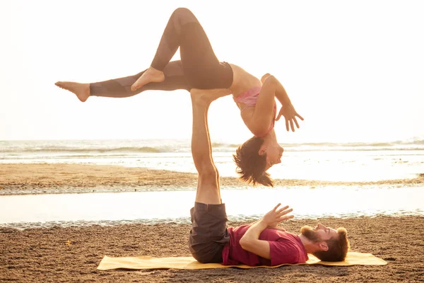 Giovane coppia sportiva ragazza e ragazzo praticano esercizi di acroyoga al tramonto sulla spiaggia — Foto Stock