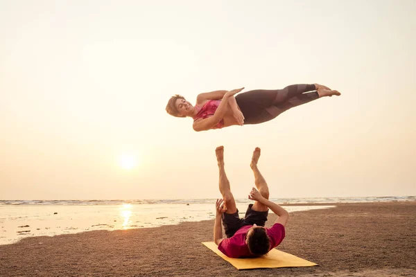 젊은 스포츠 커플 소녀와 소년은 해변에서 일몰에 아크로요가 연습을하고있습니다 — 스톡 사진