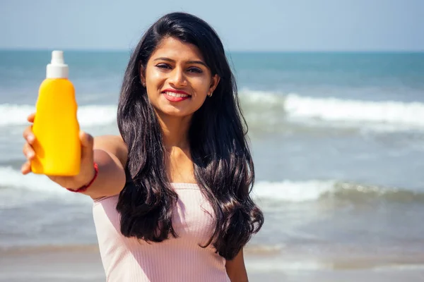 Retrato de una hermosa y sonriente sonrisa blanca como la nieve mujer india pelo negro y piel oscura en una camiseta rosa sosteniendo botella de protector solar aerosol en beach.girl disfrutando de vacaciones cuerpo spf paraíso —  Fotos de Stock