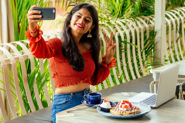 Индуистка в современном наряде сидит за столом кафе с мобильным телефоном на руках, делая селфи. Деловая женщина ест блины и работает ноутбук на летней веранде. — стоковое фото