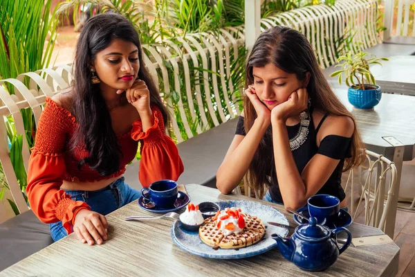 sad fashionable indian friends couple of two woman looking at cakes.friends long-awaited meeting gossip students eating pancake drinking masala tea after shopping.summer instahram lifestyle