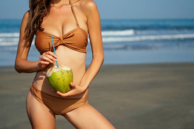 beautiful tanned girl in trendy brown bathing suit sunglasses stands with coconut in her hands on beach tropical island vacation travel swimwear new season.derox diet idea