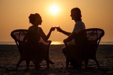 silhouette couple in love sitting on wicker chairs and clinking glasses with drinks on the beach with sunset view Valentines Day