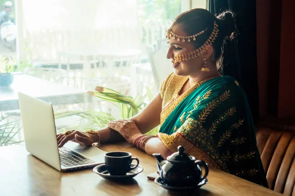 Симпатичная индийская бизнесвумен в зеленом традиционном сари использует ноутбук в офисе рядом с окном. удаленная работа и фриланс — стоковое фото