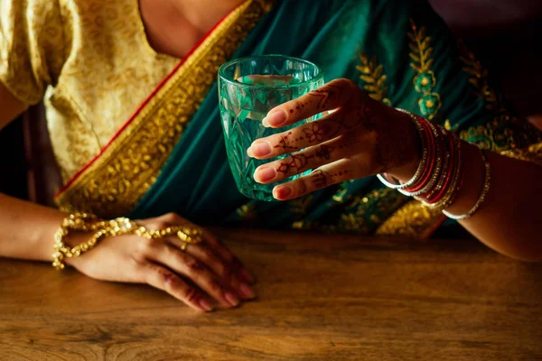 Όμορφη Ινδική Ασιατική κορίτσι σε Sari κρατώντας ένα ποτήρι σκέτο νερό, κάθεται στο σπίτι κουζίνα κοντά στο παράθυρο — Φωτογραφία Αρχείου