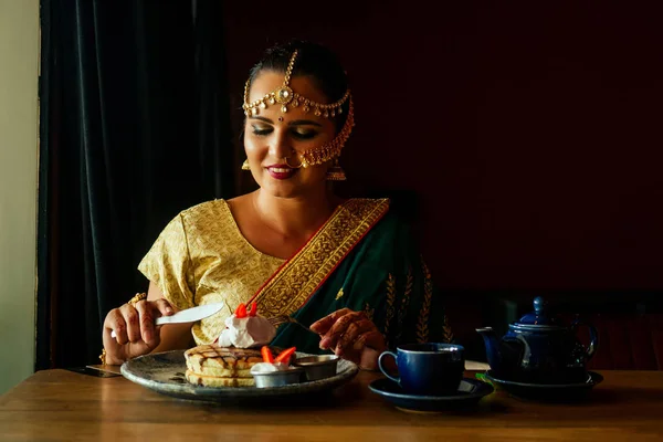 Портрет индийской женщины в зеленом сари и золотом ювелире, смотрящей на блины со взбитыми сливками и клубничным десертом, жаждущей съесть . — стоковое фото