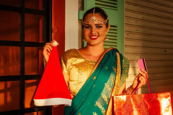 Счастливая красивая индийская женщина в традиционном платье холдинг магазин сумка и Санта-Клаус шляпа на рынке, сумки и кредитная карточка. — стоковое фото