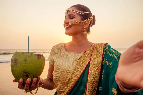 Bela mulher indiana sari moda verde, bindis beber água de noz de coco. mulher indiana perfeito maquiagem tikka, nath, pino de nariz no verão Goa praia na Índia oceano .tourist tomando selfie no telefone da câmera — Fotografia de Stock