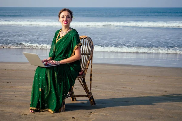 Remote working concept dream job.beautiful and young indian woman in green stylish sari, curly hairstyle, toothy white smile and flower in hair working with a laptop while sitting in a summer by sea — стоковое фото