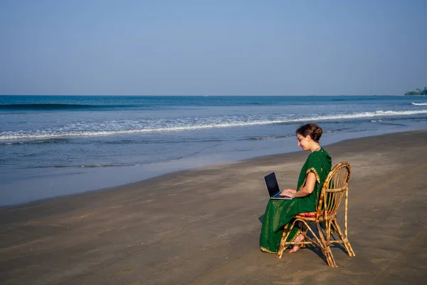 business woman india asian female freelancer sitting chair. woman in indian green stylish saree sari working on laptop and smiling smartphone.remote work dream job in the paradise coast copy space