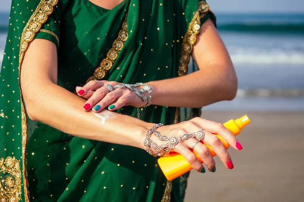 Retrato de um belo e sorridente sorriso branco-neve mulher indiana cabelo encaracolado preto e pele escura em sari verde segurando garrafa de spray protetor solar em beach.girl desfrutando spf corpo paraíso férias — Fotografia de Stock